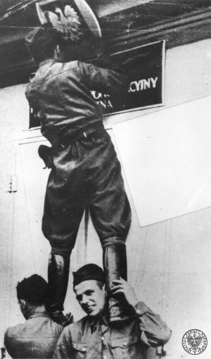 Żołnierz niemiecki stojący na barkach innych żołnierzy i usuwający tabliczkę z orłem z polskiego urzędu