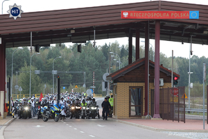 Motocykliści w zwartej grupie stoją przy przejściu granicznym. Nad nimi wiata a obok tablica z napisem: „ Rzeczpospolita Polska”.