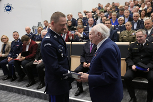 Komendant Główny Policji gen. Marek Boroń gratuluje jednemu z uczestników spotkania. W tle na widowni funkcjonariusze Policji, oficerowie Wojska Polskiego oraz goście