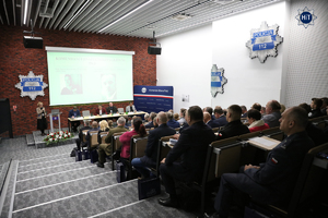 Widok na salę, w której trwa konferencja. Przy prezydialnym stole siedzą prelegenci. Za nimi duży ekran, obok policyjna gwiazda.