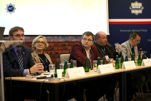 Przy prezydialnym stole siedzą prelegenci. W tle granatowy roll-up z napisem ”Komenda Główna Policji”.