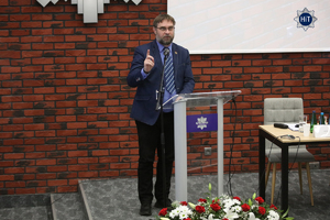 Jeden prelegentów przemawia do mikrofonu z mównicy, przy której leży wiązanka biało-czerwonych kwiatów.