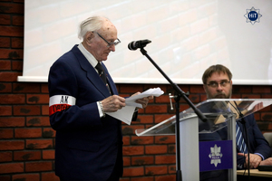 Jeden z prelegentów stoi przy mównicy i odczytuje tekst. Na przedramieniu jego marynarki widnieje biało-czerwona powstańcza opaska.