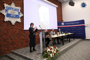 Prelegentka ubrana w mundur funkcjonariuszki Policji Państwowej przemawia do mikrofonu stojąc przy mównicy.