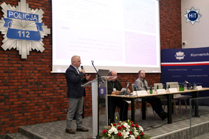Jeden prelegentów przemawia do mikrofonu z mównicy, przy której leży wiązanka biało-czerwonych kwiatów.