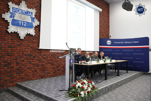 Widok na stół prezydialny, przy którym siedzą prelegenci. Na ścianie wisi policyjna gwiazda z napisem ”Policja” i numerem „112”.