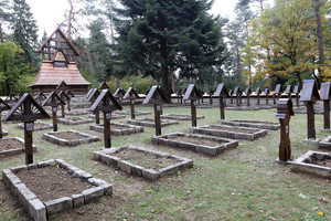 Widok na cmentarz żołnierski z jednakowymi drewnianymi krzyżami przy nagrobkach oraz prostymi mogiłami usypanymi z ziemi i obłożonymi wokół kamieniami.
