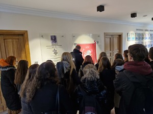 Uczniowie znajdują się w holu KGP przy ścianie, na której wisi czerwony sztandar z białym orłem oraz napisem ”Bóg, Honor i Ojczyzna”.