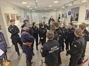 Młodzież z klasy mundurowej stoi w półokręgu holu wystawowym Wydziału Edukacji Historycznej. W środku pracownik Wydziału ubrany w mundur funkcjonariusza Policji Państwowej.