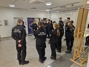 Młodzież z klasy mundurowej stoi w holu Wydziału Edukacji Historycznej. Na plecach granatowych mundurów widnieje napis: „ klasa policyjna”. W tle sztalugi i eksponaty prezentowane na wystawie.