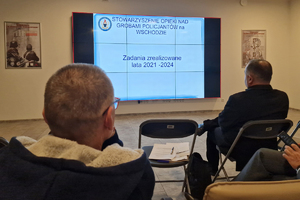 Widok na salę w budynku Wydziału Edukacji Historycznej i siedzących tam gości patrzących na duży ekran, na którym jest napis:” Stowarzyszenie nad Grobami Policjantów na Wschodzie. Zadania zrealizowane lata 2021-2024”.
