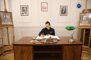 Młoda policjantka siedzi przy eleganckim drewnianym biurku w zrekonstruowanym gabinecie Komendanta Głównego Policji Państwowej.