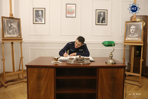 Młody policjant siedzi przy eleganckim drewnianym biurku w zrekonstruowanym gabinecie Komendanta Głównego Policji Państwowej i wpisuje się do księgi pamiątkowej.