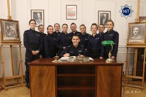 Młody policjant siedzi przy eleganckim drewnianym biurku w zrekonstruowanym gabinecie komendanta Głównego Policji Państwowej i wpisuje się do księgi pamiątkowej. Za nim stoją w zwartej grupie policjantki – reprezentantki drużyny w Futsalu. Razem pozują do zdjęcia.