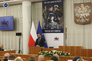Prof. Piotr Gołdyn z Uniwersytetu Kaliskiego przemawia z mównicy w Senacie. Za nim na ścianie plakat z wizerunkami funkcjonariuszek Policji i napisem ”Policja jest kobietą”.
