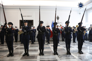 Widok na sejmowy hol z orkiestrą reprezentacyjną Policji a także z policjantkami stojącymi w szeregu i trzymającymi wyciągnięte przed siebie strzelby.