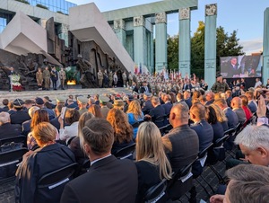 Widok na Pomnik Powstania Warszawskiego, przy którym pali się znicz. Przed pomnikiem siedzą zaproszeni goście.