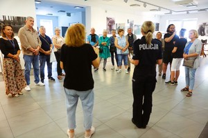 Grupa kilkunastu dorosłych osób stoi w półokręgu w holu wystawowym. Przed nimi kobieta mówiąca „językiem migowym” oraz policjantka.