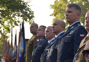 Funkcjonariusze Policji i oficerowie Wojska Polskiego stoją w szeregu , w tle sztandary wojskowe.