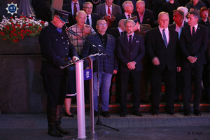Funkcjonariusz Policji ubrany w mundur Policji Państwowej czyta tekst stojąc przy mikrofonie. Za nim goście zaproszeni na uroczystość.