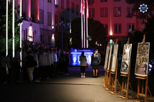 Plac przed gmachem Komendy Głównej Policji. Na środku sztandar z biało-czerwoną flagą. Na schodach budynku stoją zaproszeni goście. Jest mrok.