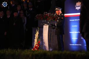 Komendant Główny Policji stoi przy mównicy. W tle baner z policyjną gwiazdą i napisem: „Komenda Główna Policji”. Jest mrok.