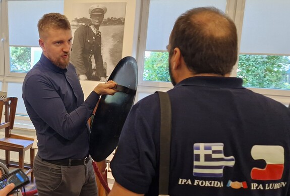 Mężczyźni oglądają zabytkowe tarcze obronne. Za nimi na ścianie wisi powiększone czarno-białe zdjęcie funkcjonariusza Policji Państwowej.