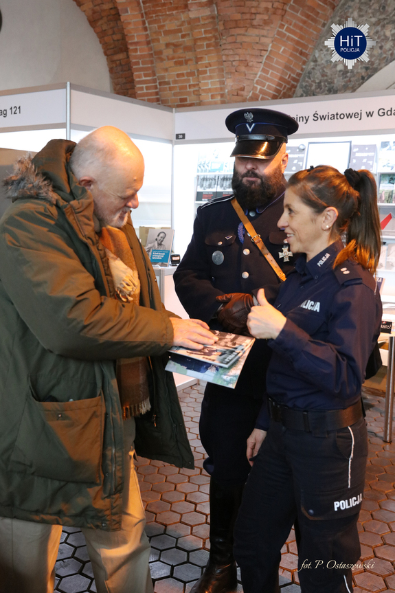 Funkcjonariuszka Policji wręcza broszurę informacyjną mężczyźnie odwiedzającemu targi. Obok niej funkcjonariusz w umundurowaniu policjanta Policji Państwowej.