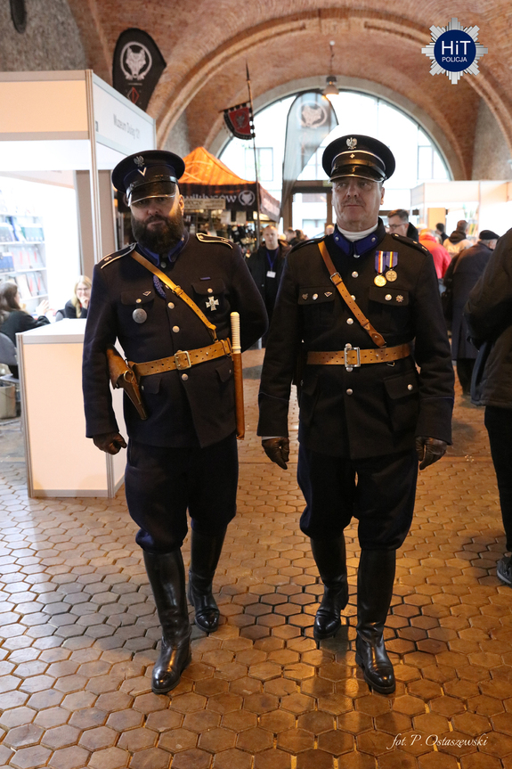 Dwóch mężczyzn w umundurowaniu funkcjonariuszy Policji Państwowej przechadza się po holu, przy którym zlokalizowane są stoiska targowe.