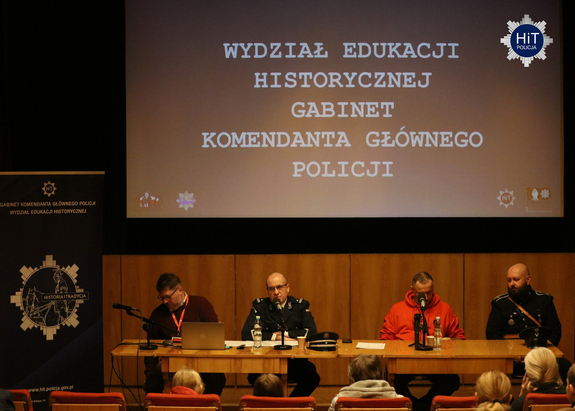 Przy stole ustawionym przed publicznością siedzi czterech mężczyzn. W tle baner z napisem: ”Wydział Edukacji Historycznej Gabinet Komendanta Głównego Policji: