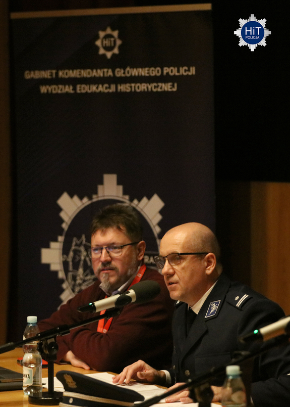 Przy stole ustawionym przed publicznością siedzi policjant, który mówi do mikrofonu. W tle baner z napisem: „Gabinet Komendanta Głównego Policji Wydział Edukacji Historycznej”.