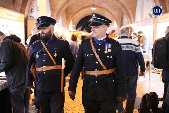 Dwóch mężczyzn ubranych w mundury funkcjonariuszy Policji Państwowej idzie wzdłuż holu, gdzie znajdują się stoiska wystawców.