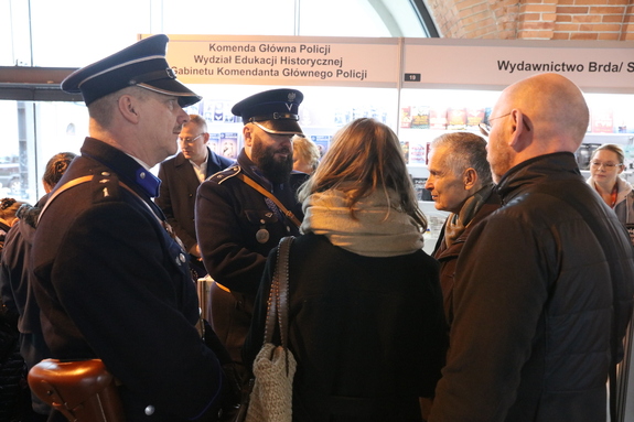 Mężczyźni w umundurowaniu funkcjonariuszy Policji Państwowej rozmawiają z odwiedzającymi targi. W tle stoisko Wydziału Edukacji Historycznej.