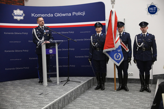 Przy mikrofonie stoi Komendant Główny Policji. Obok niego asysta honorowa Policji. W tle granatowy baner z napisem: „Komenda Główna Policji”.
