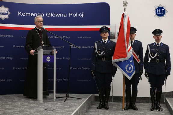 Ksiądz bisku stoi przy mikrofonie przed publicznością. Obok stoi asysta honorowa Policji.