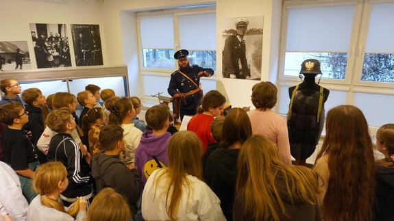 Pracownik Wydziału Edukacji Historycznej w umundurowaniu funkcjonariusza Policji Państwowej stoi przed grupą dzieci w holu wystawowym Wydziału