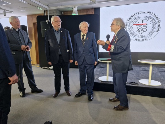 Mężczyźni stoją w półokręgu. Jeden z nich trzyma mikrofon w ręku. Na przedramieniu widnieje biało-czerwona opaska z napisem „AK”.