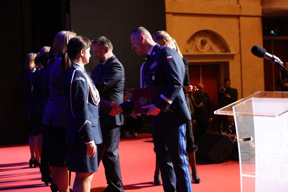 Na scenie stoją funkcjonariuszki w mundurach wyjściowych. Komendant Główny Policji oraz przedstawiciele MSWiA wręczają im odznaczenia.