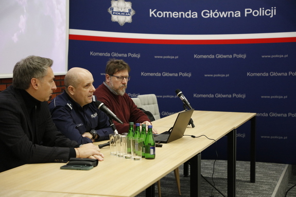Trzech mężczyzn siedzi przy stole ustawionym przed publicznością. Po prawej stronie stoi granatowy baner z biało-czerwona szarfa i napisem: ”Komenda Główna Policji”