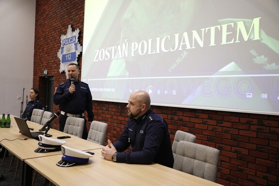Prelegenci siedzą przed słuchaczami wykładu. Na ścianie wyświetlony jest napis ostań policjantem