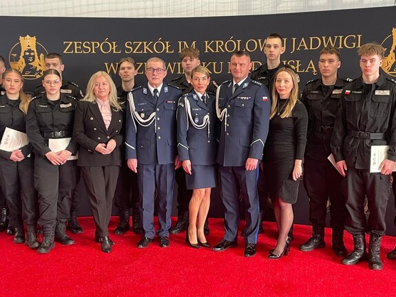 Funkcjonariusze Policji, wykładowcy a także kadeci pozują do wspólnego zdjęcia na tle baneru Zespołu Szkół im. Królowej Jadwigi w Czerwińsku nad Wisłą