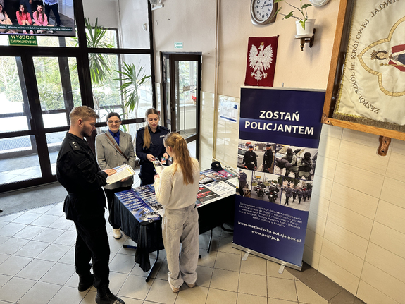 Stoisko informacyjne oraz baner z napisem: „Zostań policjantem” ustawione przy wejściu do szkoły