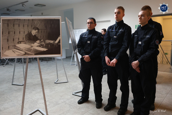 Uczniowie klasy policyjnej słuchają wykładu stojąc w holu Wydziału Edukacji Historycznej. Obok prezentowane są powiększone archiwalne fotografie.