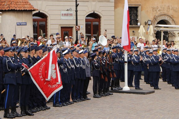 Policjanci oddają honory podczas wykonywania hymnu państwowego. Umundurowani w mundury wyjściowe prezentują haftowane sztandary policyjne.