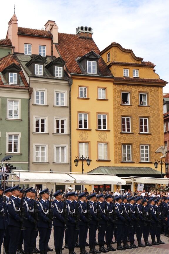 Kompania Reprezentacyjna Policji stoi w równym rzędzie na Placu Zamkowym w Warszawie. Funkcjonariusze trzymają długą broń skierowaną bagnetem ku górze. W tle zabytkowe kamienice